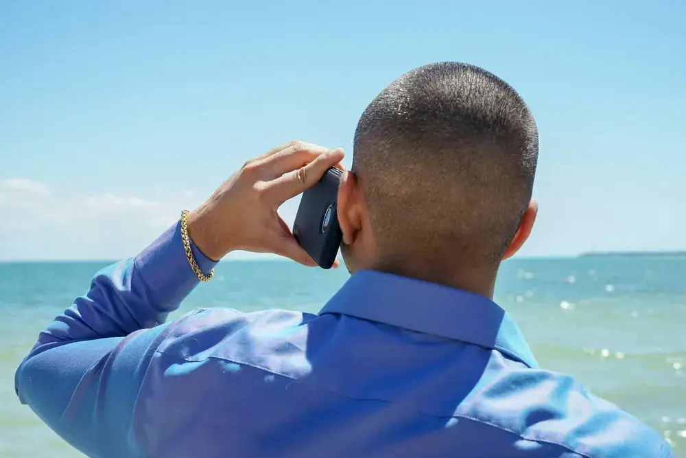 A man in a blue shirt taking a call outdoors by the water.