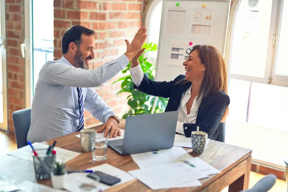 Two colleagues high-fiving over a laptop at a desk.