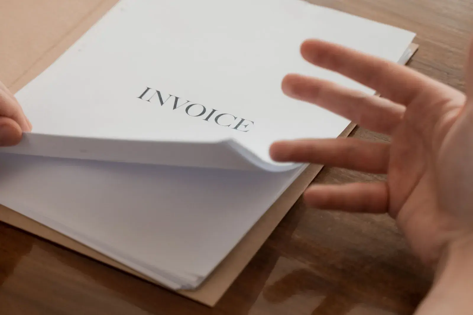 A hand turning the pages of a stack of printed invoices on a desk.
