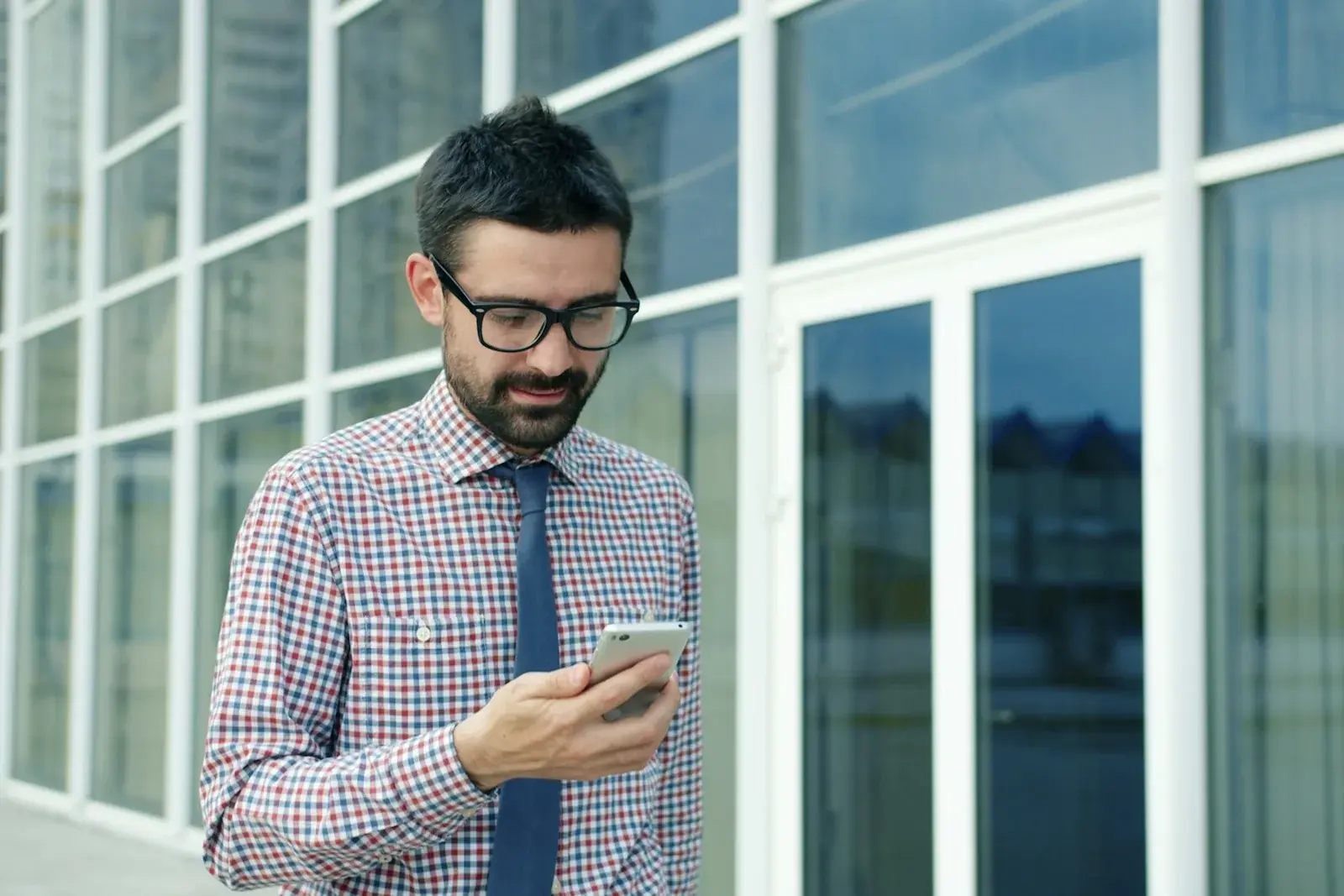 A man in a checked shirt and tie looking at his phone screen outside an office building.