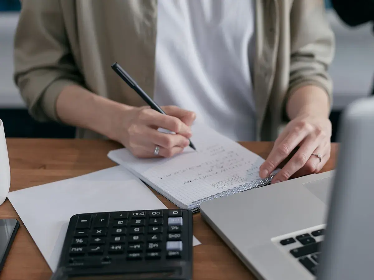 Someone working out figures in a notebook with a calculator and laptop.