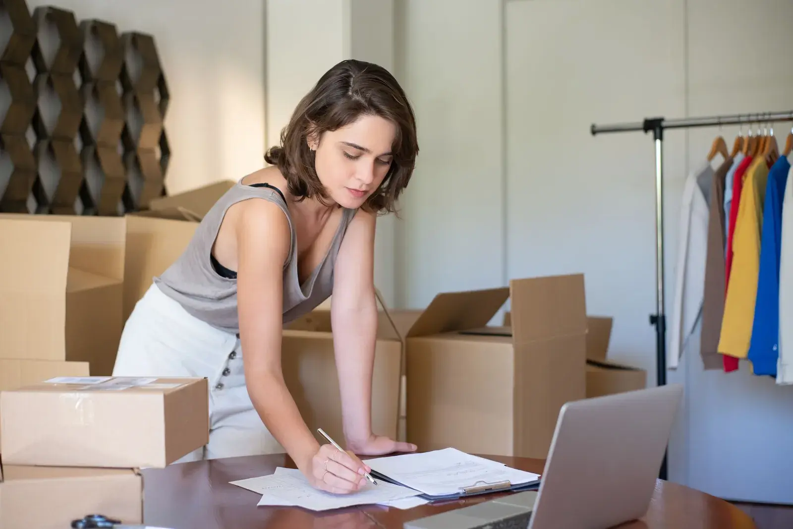 A woman checking an order sheet beside packed boxes and a rail of clothing.