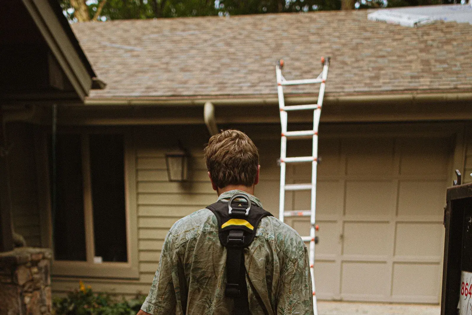 A roofer carrying harness equipment toward a house with a ladder set against the roofline.