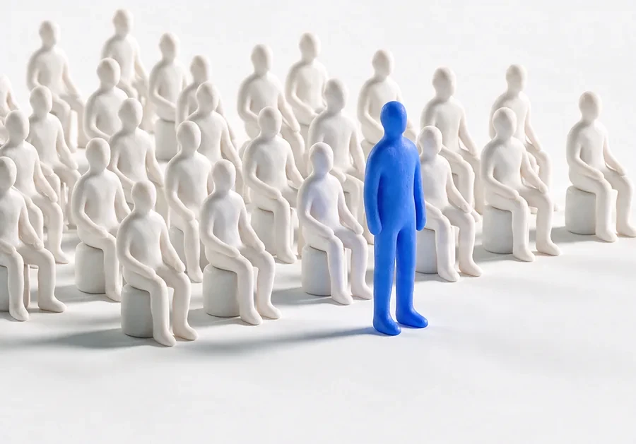A single blue figure standing among rows of seated white figures.
