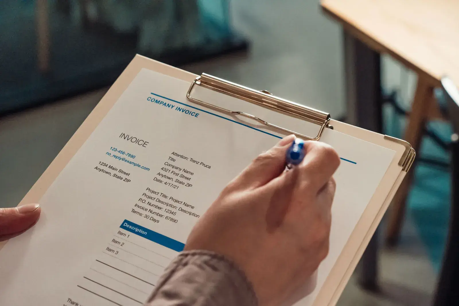 A hand holding a company invoice on a clipboard with a pen.