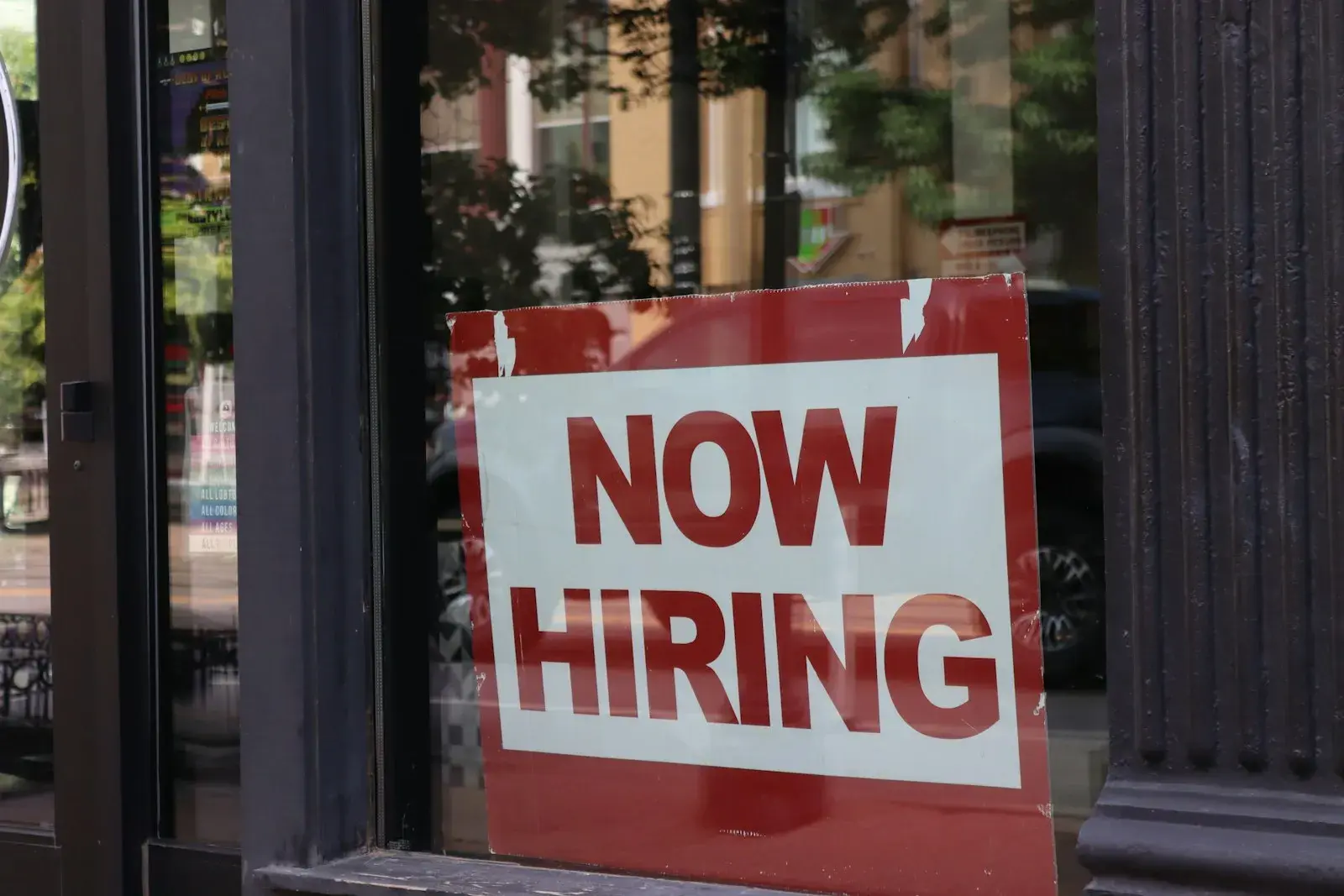 A weathered NOW HIRING sign propped in a shop window, with the street reflected in the glass.