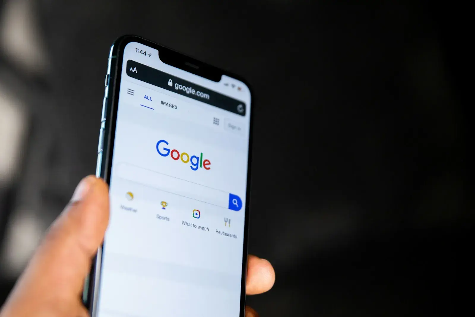 A hand holding a phone with the Google search page open and the search box still empty.